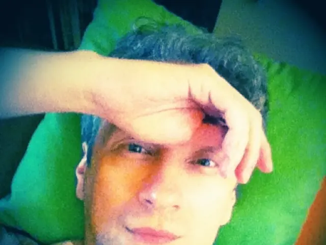 In this picture there is a man who is wearing t-shirt and lying on the bed. Behind him I can see...