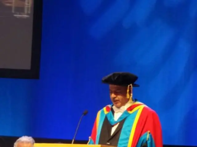 In this image we can see a man is standing. He is wearing a graduation gown and cap. In front of...