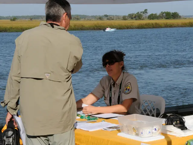 The picture is clicked in front of a lake & there is an officer sat on chair in front of table...