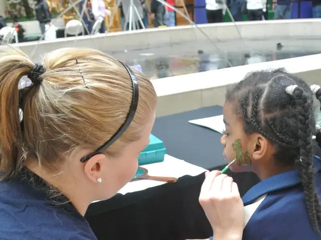 In this image we can see a girl and a woman is sitting. The woman is drawing tattoo on the cheek of...